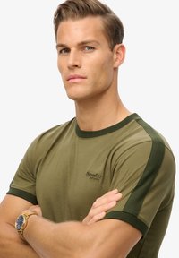 Camiseta verde oliva de manga corta, con acentos en verde más oscuro en los hombros y un pequeño logo en el lado izquierdo del pecho. Textura suave.