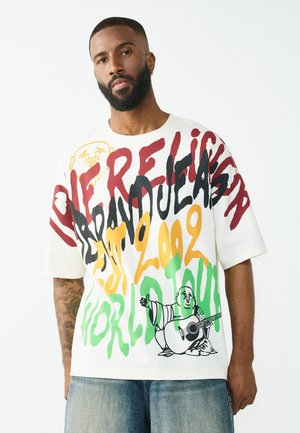 Man met baard die een witte oversized t-shirt draagt met kleurrijke graffiti tekst en een illustratie van Boeddha die gitaar speelt, gecombineerd met lichtblauwe jeans.