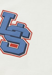 Vit t-shirt med en stor, texturerad blå och marinblå grafik med röd kontur, som visar stiliserade bokstäver "L" och "S."
