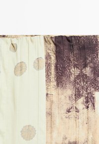 Rideau avec deux panneaux : gauche crème avec des motifs circulaires pâles, droite beige et marron foncé avec des dessins floraux et géométriques complexes.