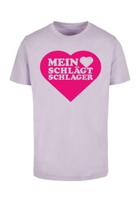 Hele lilla puuvillane T-särk, millel on suur roosa südame graafika ja tekst "MEIN ♥ SCHLÄGT SCHLAGER" rasvastes valgetes tähtedes.