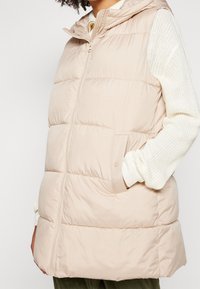 Beige prošívaná vesta s kapucí, s předním zipem, bočními kapsami a lehkým prošívaným designem. Pod ní jsou texturované pletené rukávy.