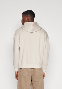 Crèmekleurige hoodie met een ontspannen pasvorm, ribgebreide manchetten en zoom; gecombineerd met lichtbeige corduroy broeken, gezien vanuit de achterkant.