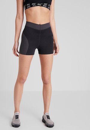 Nainen, joka pukeutunut mustiin korkeavyötäröisiin urheilushortseihin, mustaan Nike-urheiluliiviin ja harmaisiin slip-on-tossuihin, seisoo yksinkertaisella taustalla.