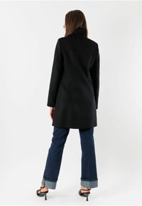 Cappotto nero su misura con colletto alto, maniche lunghe e tasche laterali; abbinato a jeans in denim scuri e sandali neri con tacco.