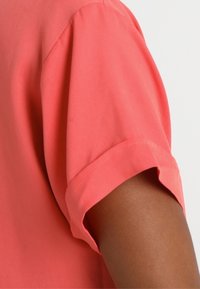 Camisa coral de manga corta con puños remangados, que presenta una textura suave de algodón y un diseño ajustado, enfatizando una apariencia estructurada.