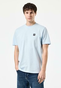 Lichtblauw katoenen T-shirt met een ronde hals, korte mouwen en een zwart vierkant logo-opdruk op de linkerkant van de borst. Draagt met donkerblauwe jeans.