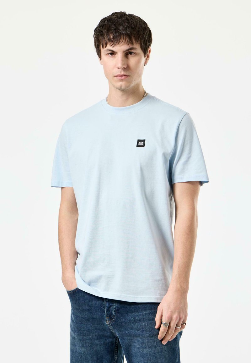 Lichtblauw katoenen T-shirt met een ronde hals, korte mouwen en een zwart vierkant logo-opdruk op de linkerkant van de borst. Draagt met donkerblauwe jeans.