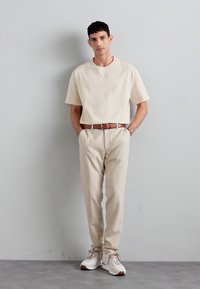 Beige short-sleeve t-shirt, cream textured pants with pockets, brown leather belt, and white sneakers with brown accents. Standing pose.
