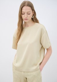 Beige kortærmet top lavet af glat, blødt stof med rund halsudskæring og afslappet pasform. Stilmæssigt kombineret med lyse bukser, der viser en subtil tekstur.