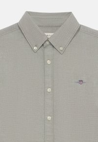Camisa abotonada gris claro con cuello puntiagudo, con botones de color crema y un pequeño logotipo bordado en el lado izquierdo del pecho.