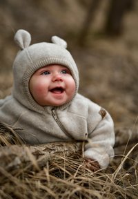 Hellbeige Fleecejacke mit Reißverschluss, mit Bärenohren auf der Kapuze. Der Hintergrund besteht aus trockenem Gras und Zweigen. Helle blaue Augen sind sichtbar.