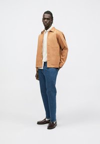 Ung mann som står, iført en beige jakke over en hvit rutete skjorte, blå jeans og brune loafers, mot en ensfarget lys grå bakgrunn.