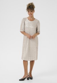 Paillettenkleid in Hellgold mit kurzen Puffärmeln und einem runden Ausschnitt. Kombiniert mit schwarzen Slingback-Absätzen. Glatte Textur, knielang.
