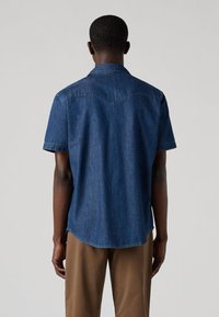 Πουκάμισο από τζιν με κοντά μανίκια σε σκούρο μπλε χρώμα, με μυτερό γιακά, ορατές ραφές και κυρτή κάτω ραφή. Το ύφασμα φαίνεται λείο με ελαφριά γυαλάδα.