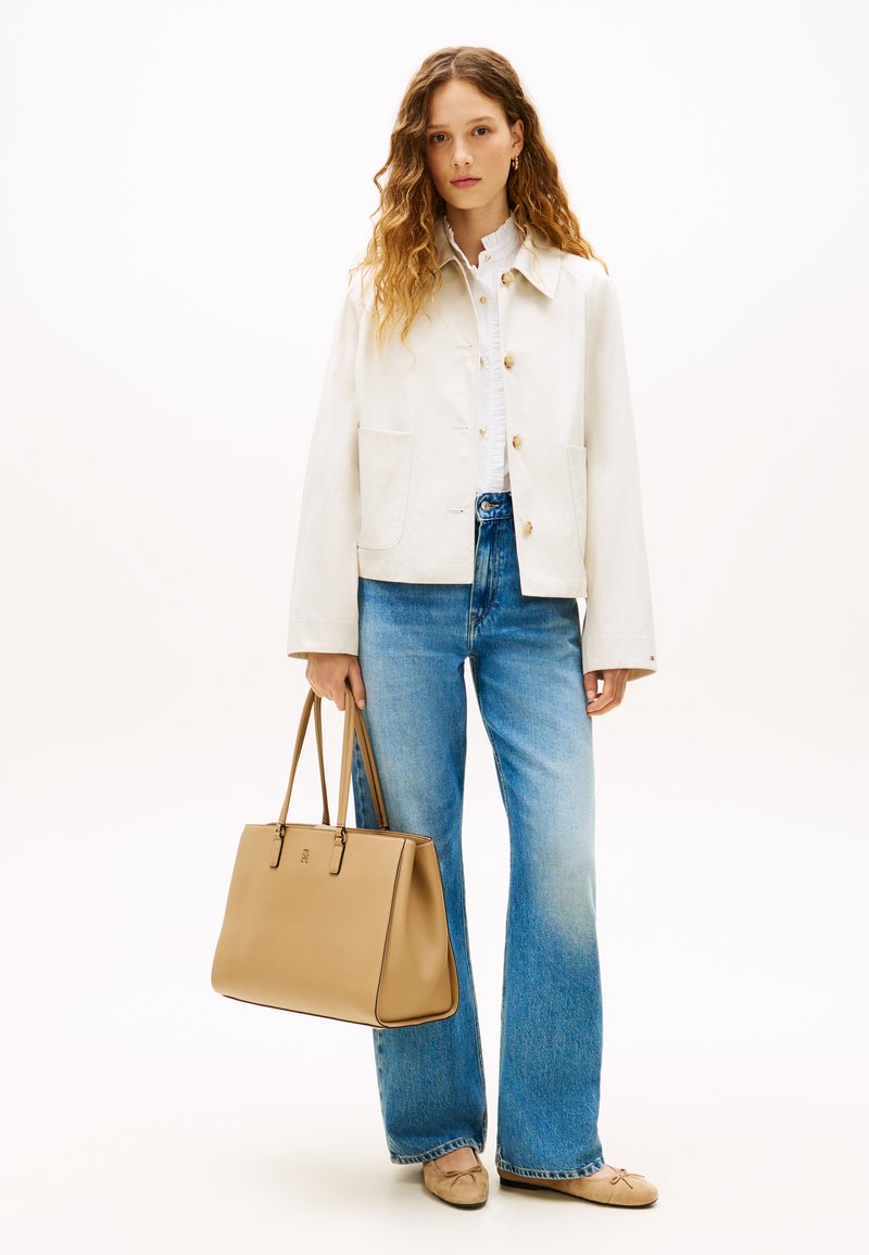 Γυναίκα με σγουρά μαλλιά που φοράει λευκό μπουφάν και μπλε τζιν, κρατώντας μια μπεζ τσάντα ώμου, στέκεται μπροστά σε ένα απλό λευκό φόντο.