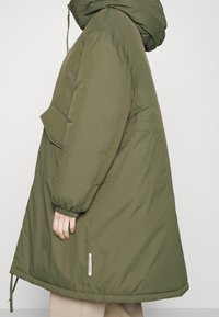 Olivgrön isolerad parkas med huva, elastiska ärmslut, ficka på sidan och dragsko i nederkant för justerbar passform. Enkel, matt textur.