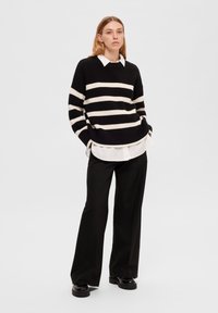 Maglione lavorato a maglia a righe bianche e nere, dalla vestibilità oversized, con scollatura rotonda, indossato sopra una camicia bianca con colletto, abbinato a pantaloni neri a gamba larga.