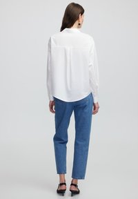Weißes Langarmhemd mit abgerundetem Saum und Rückenpasse, kombiniert mit hellblauen Jeans und schwarzen Absatzsandalen.