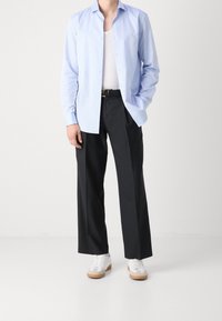 Světlomodrá košile na knoflíky, černé plisované kalhoty a bílé tenisky s béžovými detaily. Košile je mírně rozhalená, pod ní je vidět bílý tílko.