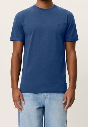 Homme portant un t-shirt bleu uni à manches courtes et un jean bleu clair, debout devant un fond uni.