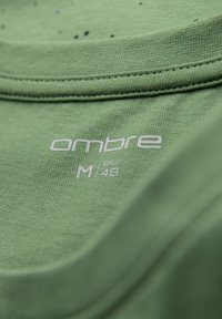 Zelené bavlněné tričko s kulatým výstřihem. Na látce se nachází název značky "ombre" a velikost "M 48" v bílém písmu. Jemná textura.