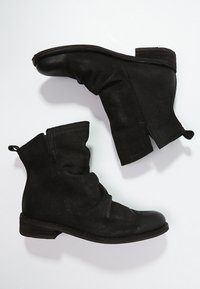 Un par de botas de tobillo negras texturizadas con tacones bajos apilados y tiradores, posicionadas sobre un fondo blanco.