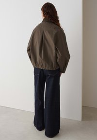 Olijfgroene bomberjack met een geplooide achterkant en geribbelde kraag, gecombineerd met brede, donkerblauwe denim jeans. Matte stof met minimale details.