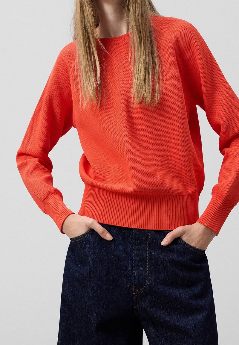 Rode gebreide trui met geribde manchetten en zoom, gecombineerd met donkerblauwe denimbroek. De stof heeft een gladde textuur en het ontwerp is eenvoudig.