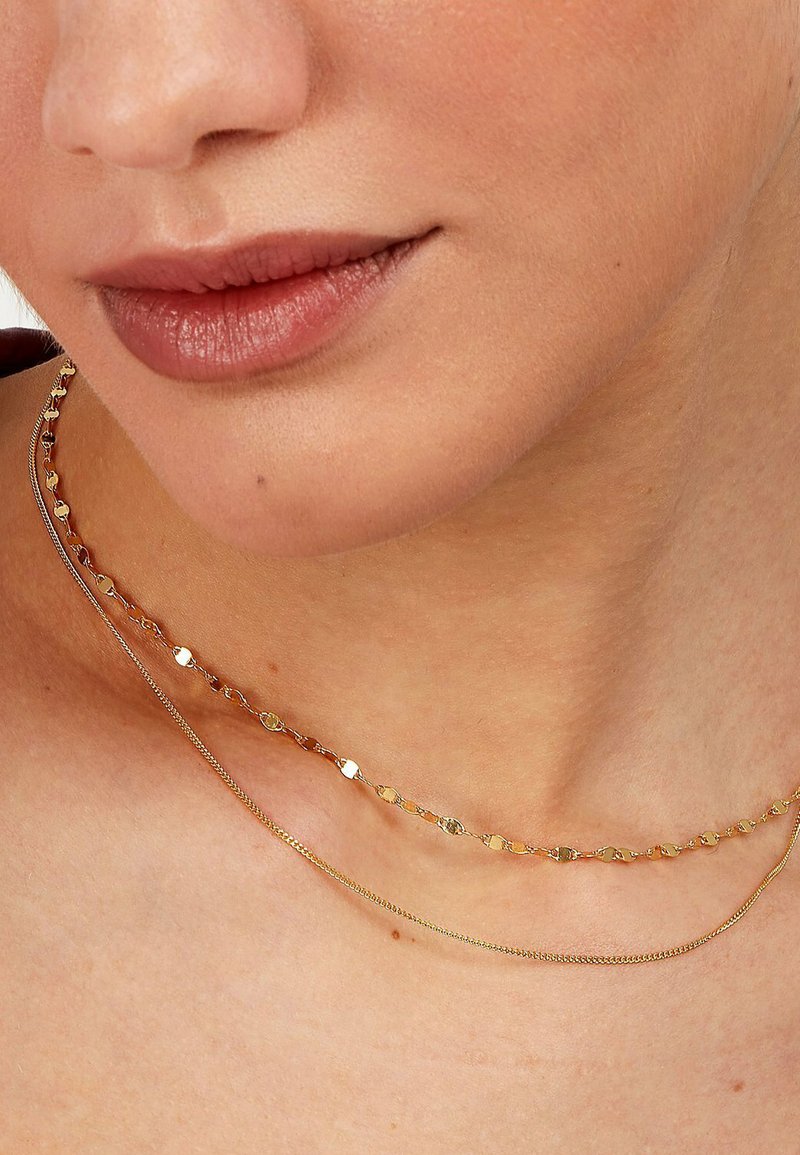 Gold- und silberne geschichtete Halsketten mit runden und ovalen Gliedern, die auf der oberen Kette strukturierte Akzente und auf der unteren eine glatte Oberfläche aufweisen.