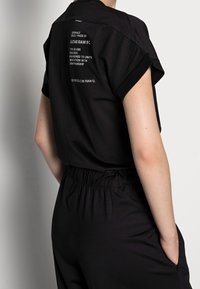 Svart jumpsuit med korta ärmar, elastisk midja och detalj med branding i vit text på ryggen. Mjuk tygkvalitet med en avslappnad passform.