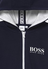 Navy zip-up huppari, jossa on valkoinen vuori. Edessä on valkoisilla brodeerattu "BOSS" ja "HUGO BOSS", sekä erottuva sisäkaulustarra.