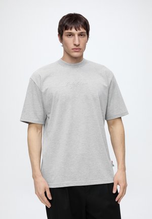 Ung mand med kort, mørkt hår iført en lysegrå kortærmet T-shirt med diskret broderet tekst på brystet og sorte bukser.