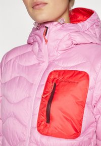 Roze gewatteerde jas met een quiltstructuur, een rode ritszak en een verstelbare capuchon met contrasterende trekkoorden.