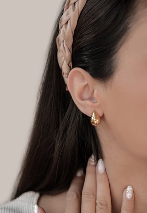 Gold teardrop earrings with a polished finish, worn on a model with dark hair, paired with a pale braided headband.