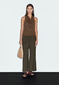 Top senza maniche in maglia marrone, pantaloni larghi oliva, borsa a spalla beige e sandali scuri con suola in legno, posizionati su uno sfondo chiaro.