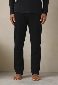 Zwarte loungebroek met een ontspannen pasvorm, gemaakt van een gladde stof; heeft een eenvoudig ontwerp en een elastische tailleband, getoond met blote voeten.
