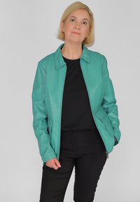 Teal Lederjacke mit Kragen, Frontreißverschluss und Seitentaschen; getragen über einem schwarzen Oberteil, kombiniert mit schwarzen Hosen.