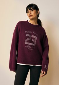 Burgundin neulepaita, jossa on ribattu helma ja valkoinen teksti "Madison Avenue 23 Athletic Club Manhattan" sekä numero "23" hihassa.
