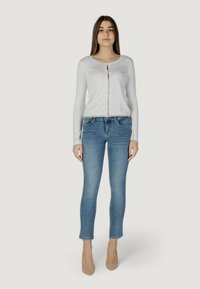 Maglietta bianca a maniche lunghe con chiusura a bottoni, abbinata a jeans azzurri chiari. I jeans hanno una vestibilità slim e un leggero sfilacciamento alle caviglie.