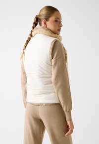 Witte gewatteerde bodywarmer met een nepbontkraag, gecombineerd met een zandkleurige gebreide trui. De bodywarmer heeft een getailleerd ontwerp en een gladde textuur.