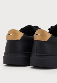 Black sneakers with thick soles and tan leather heel accents featuring small stitched flag details, shown from the back on a white background.