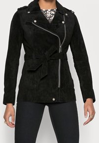 Svart mockajacka med bälte vid midjan, asymmetrisk dragkedja och axelremmar. Har en mjuk textur och en figurnära design.