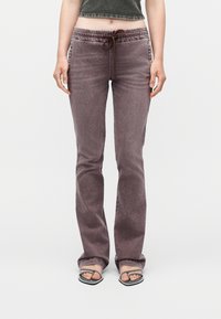 Γυναίκα που φοράει ψηλόμεσα, ξεβαμμένα, μωβ παντελόνια με ίσια γραμμή και κορδόνι στη μέση, συνοδευόμενα από μαύρες πέδιλες με ανοιχτό δάχτυλο και λεπτά λουράκια.