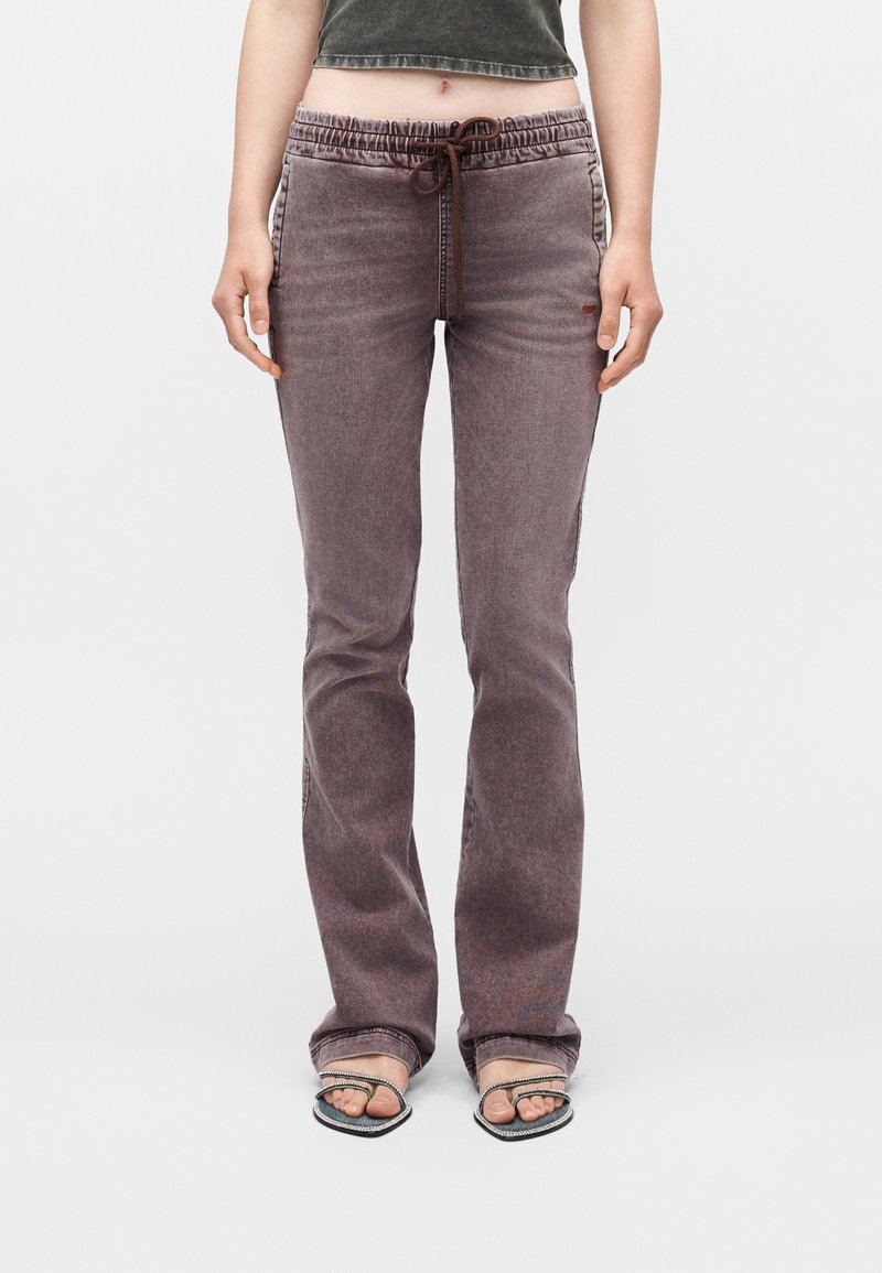 Γυναίκα που φοράει ψηλόμεσα, ξεβαμμένα, μωβ παντελόνια με ίσια γραμμή και κορδόνι στη μέση, συνοδευόμενα από μαύρες πέδιλες με ανοιχτό δάχτυλο και λεπτά λουράκια.