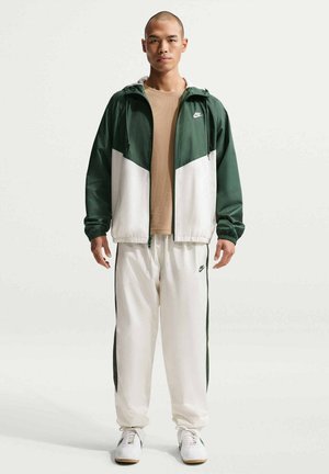 Man met een groen-wit Nike jack, een beige overhemd, witte Nike trainingsbroek en witte sneakers, staand met gezicht naar voren tegen een effen achtergrond.