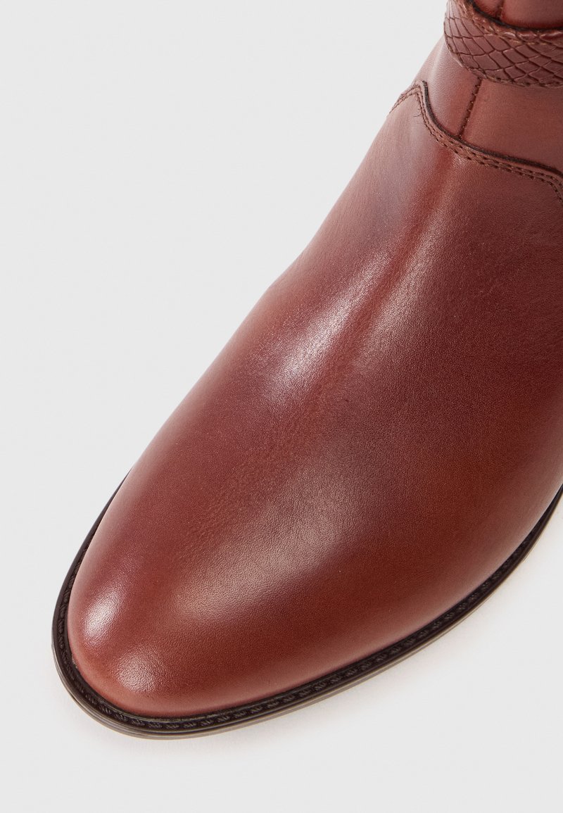 Stivale in pelle marrone alla caviglia con una finitura liscia, punta affusolata e dettaglio intrecciato vicino alla parte superiore. Presenta una cucitura a contrasto attorno alla suola.
