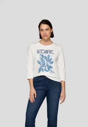 Weißes Baumwoll-T-Shirt mit blauem grafischem Design, das das Wort "ICONIC" und blaue Blattmuster zeigt, kombiniert mit dunkelblauen Jeans.