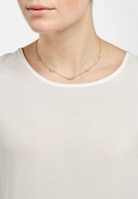 Gouden ketting met vijf kleine, duidelijke versieringen. Draagt boven een eenvoudige witte shirt met een ronde hals.