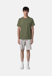 T-shirt verde oliva a maniche corte con tasca sul petto, abbinato a pantaloni corti grigi chiari. Sneakers bianche e calzini alla caviglia completano il look.